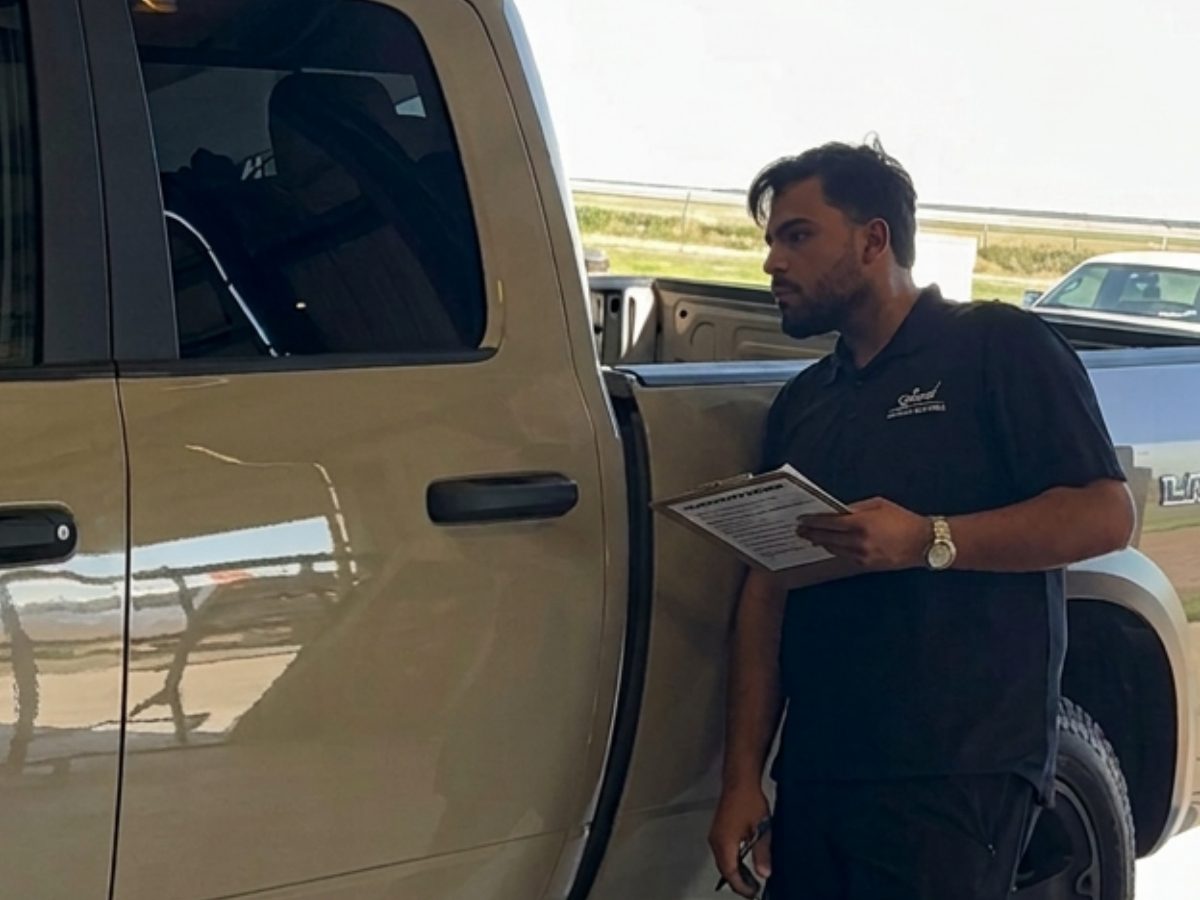 A technician beside a tan pickup, filling out an inspection sheet on a clipboard