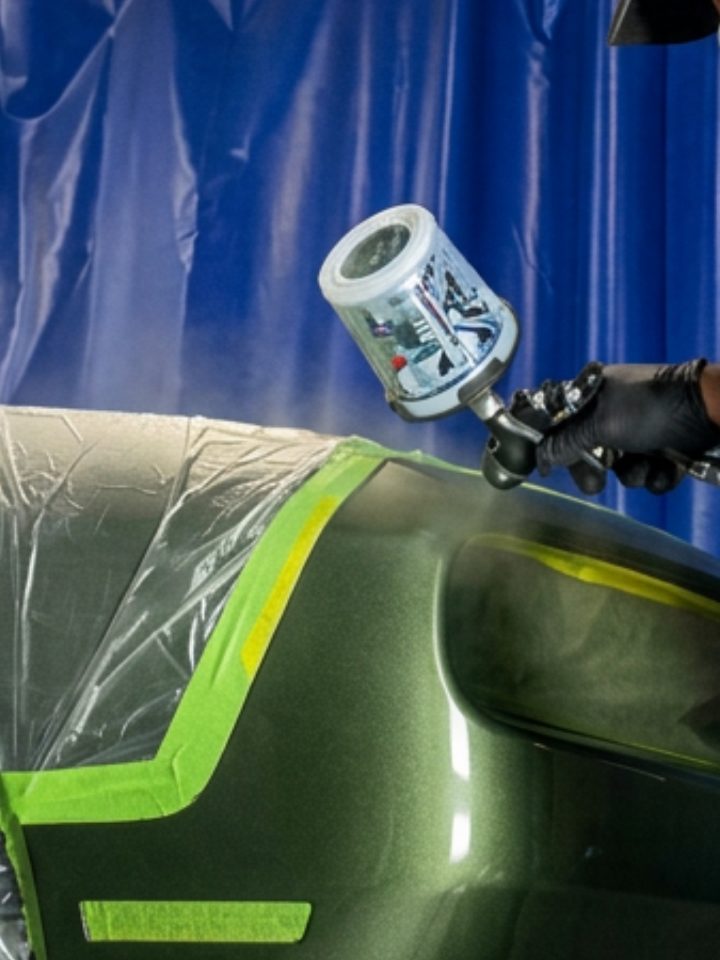 A technician spraying paint onto a masked green bumper inside the paint booth