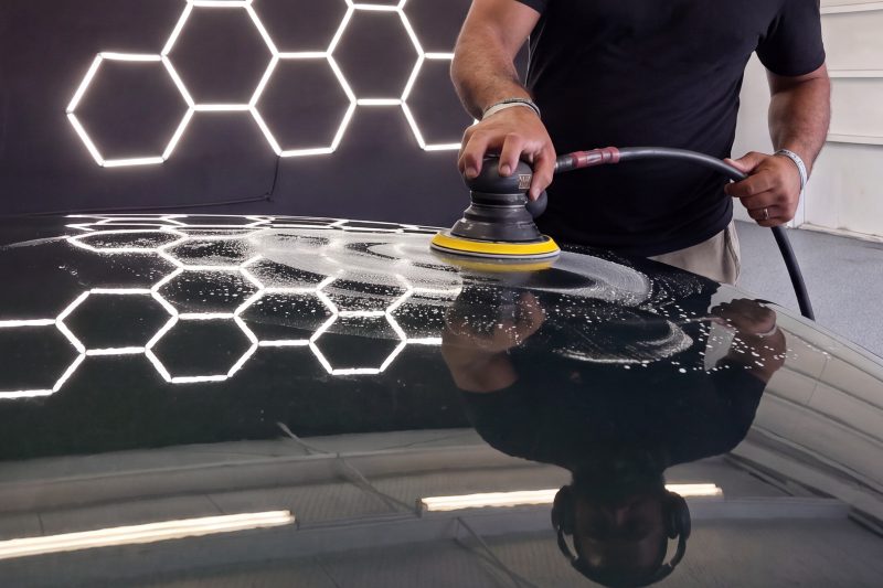 A polisher working compound across a black hood, the hexagon lights reflected in the paint